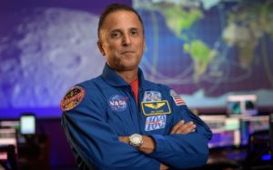 Retrato de Joe Acabá, nuevo jefe de astronautas de la NASA. Acabá, de pelo corto y oscuro, ojos oscuros y luciendo una media sonrisa, va vestido con el mono azul de los astronautas y cruza los brazos. De fondo se ven grandes pantallas con imágenes de la Luna y un mapa de la Tierra.